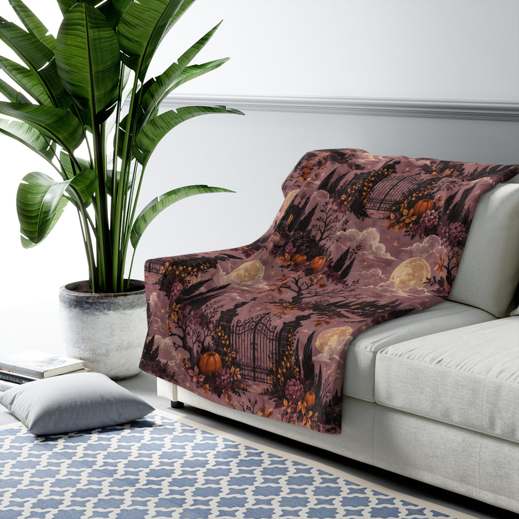 A mauve-toned throw blanket with a spooky graveyard and pumpkin pattern is draped over a light gray sofa in a styled living room setting with a potted plant and patterned rug.