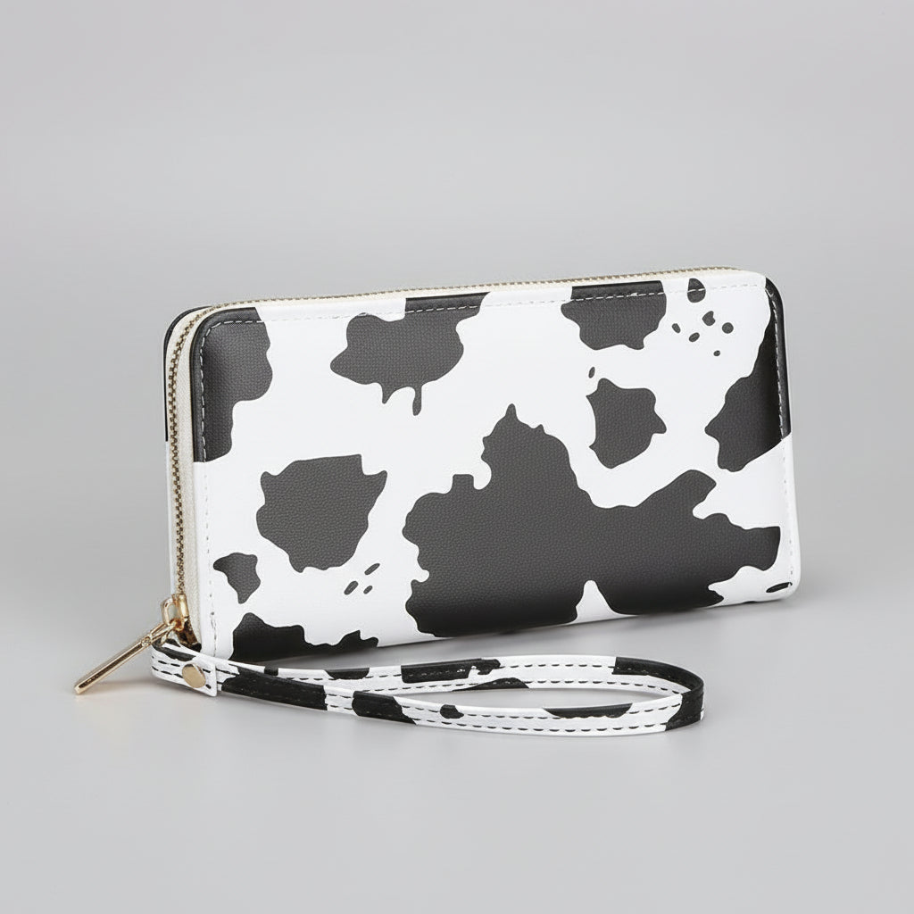 Hand holding a cow print wallet on a wooden surface with a cup and flowers in the background.